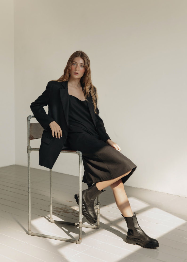 K&ouml;nnte beinhalten: Eine Person tr&auml;gt einen schwarzen Blazer, ein Kleid und Stiefel. Der Blazer hat einen klassischen Schnitt, und das Kleid hat einen einfachen Ausschnitt. Die Stiefel sind schwarz mit einer klobigen Sohle. Die Person sitzt auf einem Metallstuhl in einem Raum mit wei&szlig;en W&auml;nden und Holzboden.
