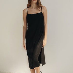 K&ouml;nnte beinhalten: Ein schwarzes Midikleid mit d&uuml;nnen Tr&auml;gern und einem Seitenschlitz. Das Kleid wird mit schwarzen Riemchensandalen kombiniert. Das Model steht vor einer wei&szlig;en Wand.