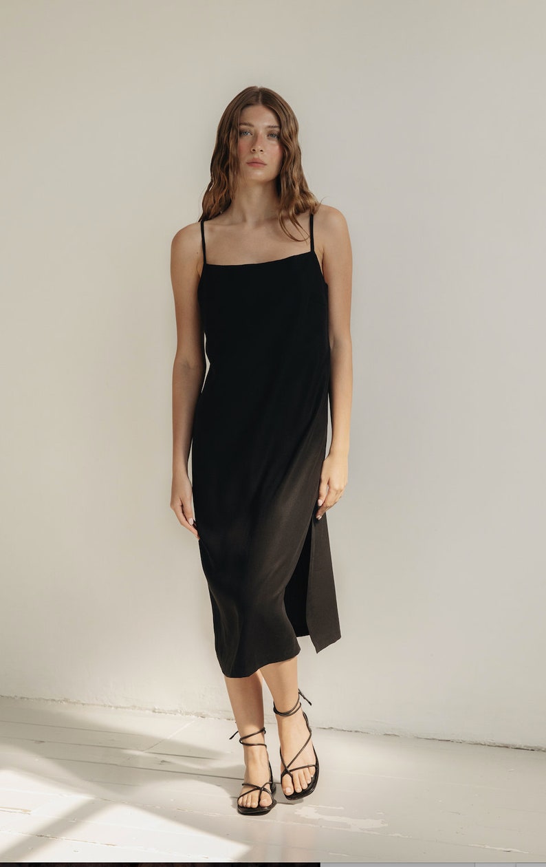 K&ouml;nnte beinhalten: Ein schwarzes Midikleid mit d&uuml;nnen Tr&auml;gern und einem Seitenschlitz. Das Kleid wird mit schwarzen Riemchensandalen kombiniert. Das Model steht vor einer wei&szlig;en Wand.