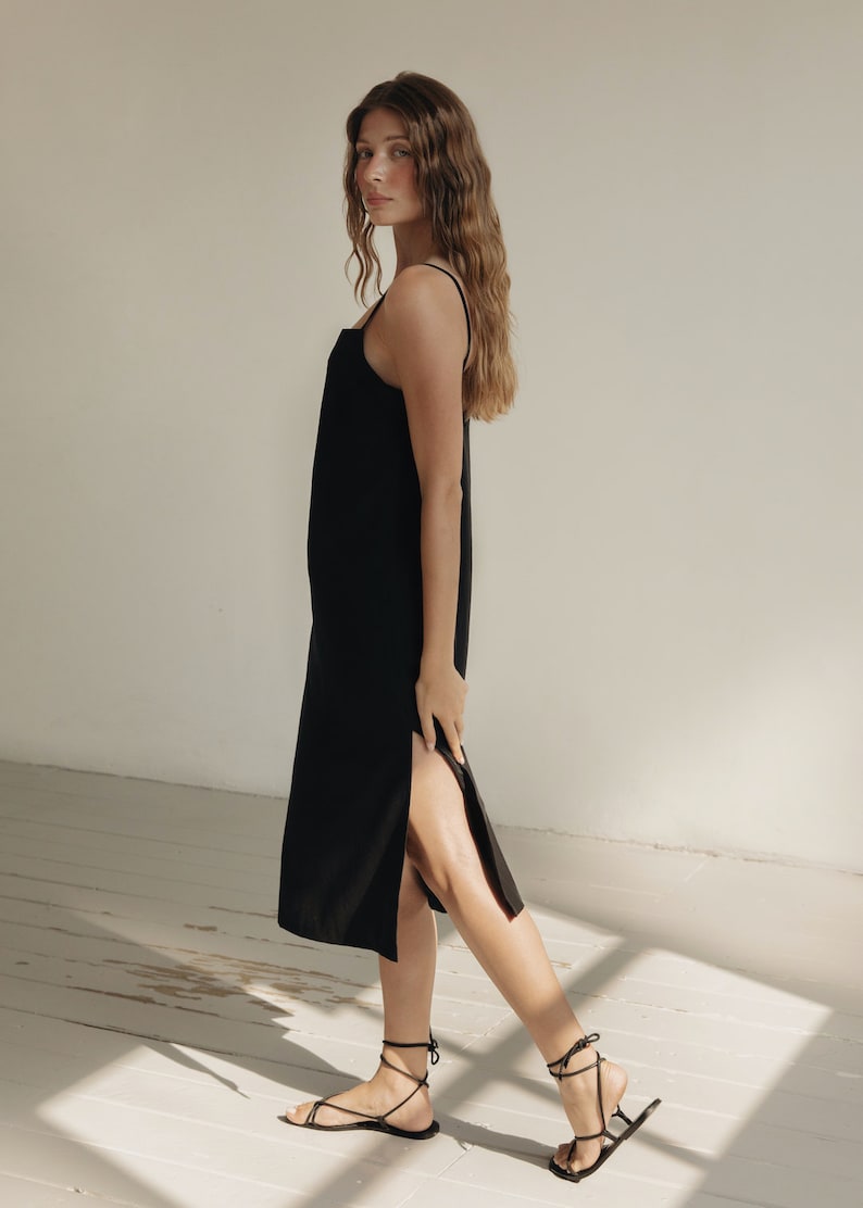 K&ouml;nnte beinhalten: Schwarzes Midikleid mit d&uuml;nnen Tr&auml;gern und Seitenschlitz. Das Kleid wird mit schwarzen Riemchensandalen kombiniert. Das Model steht in einem Raum mit wei&szlig;en W&auml;nden und einem wei&szlig;en Holzboden.
