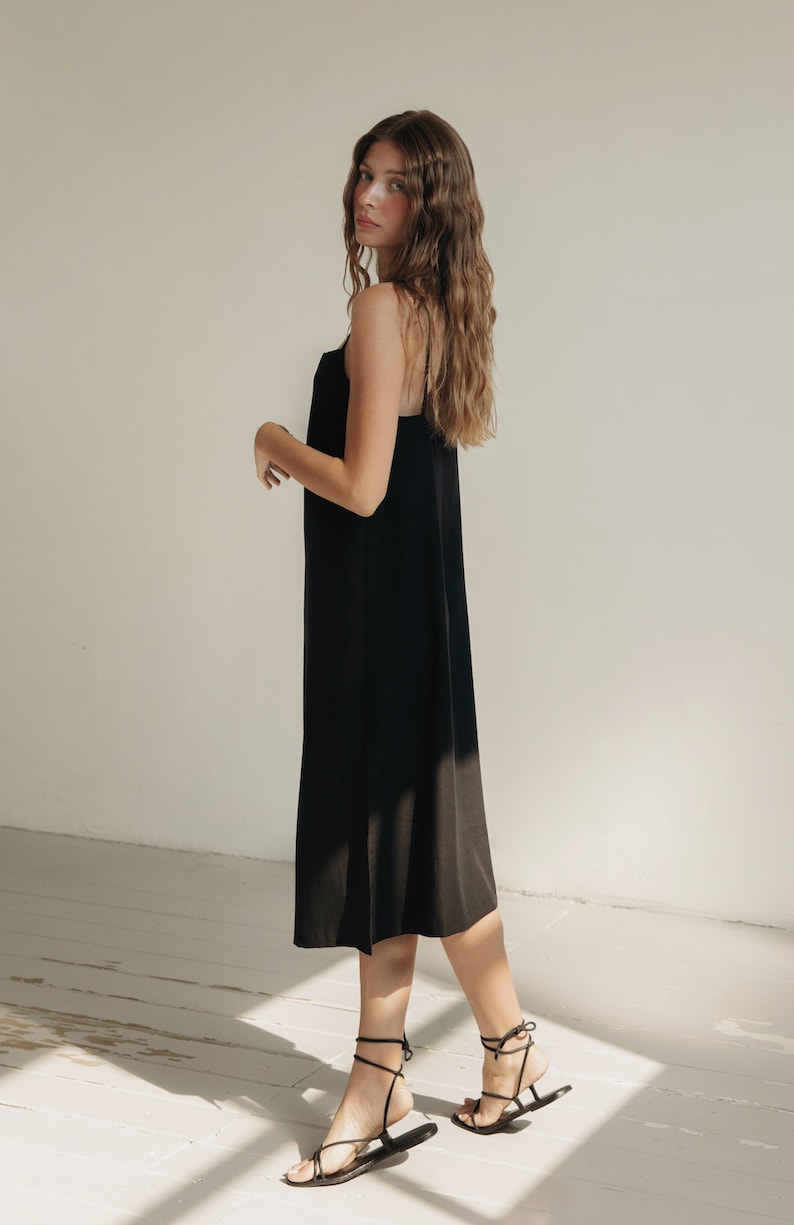 K&ouml;nnte beinhalten: Ein schwarzes Midikleid mit Spaghettitr&auml;gern. Das Kleid wird mit schwarzen Riemchensandalen kombiniert. Der Hintergrund ist ein Raum mit wei&szlig;en W&auml;nden und Holzboden.