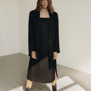 K&ouml;nnte beinhalten: Eine Person tr&auml;gt einen schwarzen Blazer &uuml;ber einem schwarzen Kleid mit quadratischem Ausschnitt und schwarzen Stiefeletten. Das Kleid hat einen Seitenschlitz. Das Outfit befindet sich vor einem wei&szlig;en Hintergrund mit nat&uuml;rlichem Licht.
