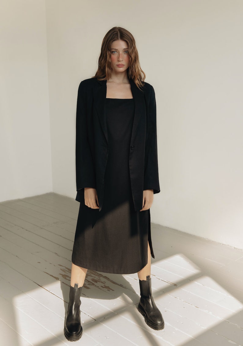 K&ouml;nnte beinhalten: Eine Person tr&auml;gt einen schwarzen Blazer &uuml;ber einem schwarzen Kleid mit quadratischem Ausschnitt und schwarzen Stiefeletten. Das Kleid hat einen Seitenschlitz. Das Outfit befindet sich vor einem wei&szlig;en Hintergrund mit nat&uuml;rlichem Licht.