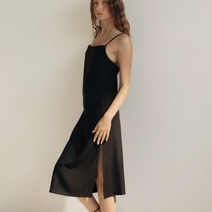 K&ouml;nnte beinhalten: Ein schwarzes Midikleid mit d&uuml;nnen Tr&auml;gern und einem Seitenschlitz. Das Kleid ist aus leichtem Stoff gefertigt und hat ein schlichtes, elegantes Design. Das Model tr&auml;gt schwarze Riemchensandalen.