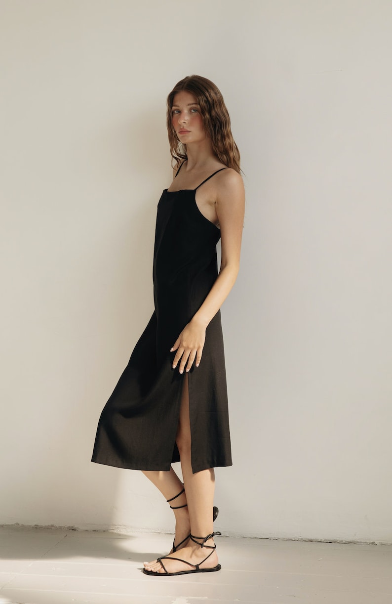 K&ouml;nnte beinhalten: Ein schwarzes Midikleid mit d&uuml;nnen Tr&auml;gern und einem Seitenschlitz. Das Kleid ist aus leichtem Stoff gefertigt und hat ein schlichtes, elegantes Design. Das Model tr&auml;gt schwarze Riemchensandalen.