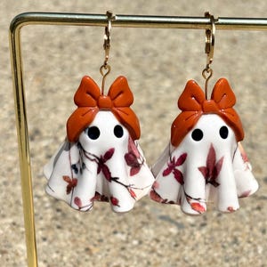 May include: A pair of dangle earrings featuring white ghost-shaped charms with black circular eyes, topped with burnt orange bows. The ghost bodies are decorated with a pattern of dark red and brown autumn leaves and branches, hanging from gold-toned hooks.