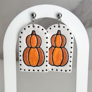 May include: A pair of ceramic earrings featuring a pumpkin design. The earrings are white with orange and brown pumpkin illustrations and black dot accents. They hang from silver-colored posts and are displayed on a white stand with a wooden base.