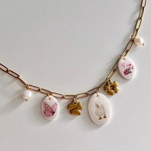 May include: Gold-tone chain necklace with various charms. Charms include a pink butterfly, a flower, a duck, and a floral design. The necklace also features a pearl accent. The charms are oval-shaped and white with pink and gold accents.