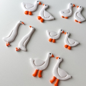 May include: White goose-shaped embellishments with orange feet and beaks are arranged on a white surface. The geese are in various poses, some facing each other, others standing alone. These decorative elements could be used for crafts or other creative projects.