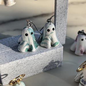 May include: A pair of drop earrings featuring small, ghost-shaped charms made of white ceramic with swirling teal and green marbled patterns. Each ghost has two small black dots for eyes and is attached to a silver-toned metal earring hook.