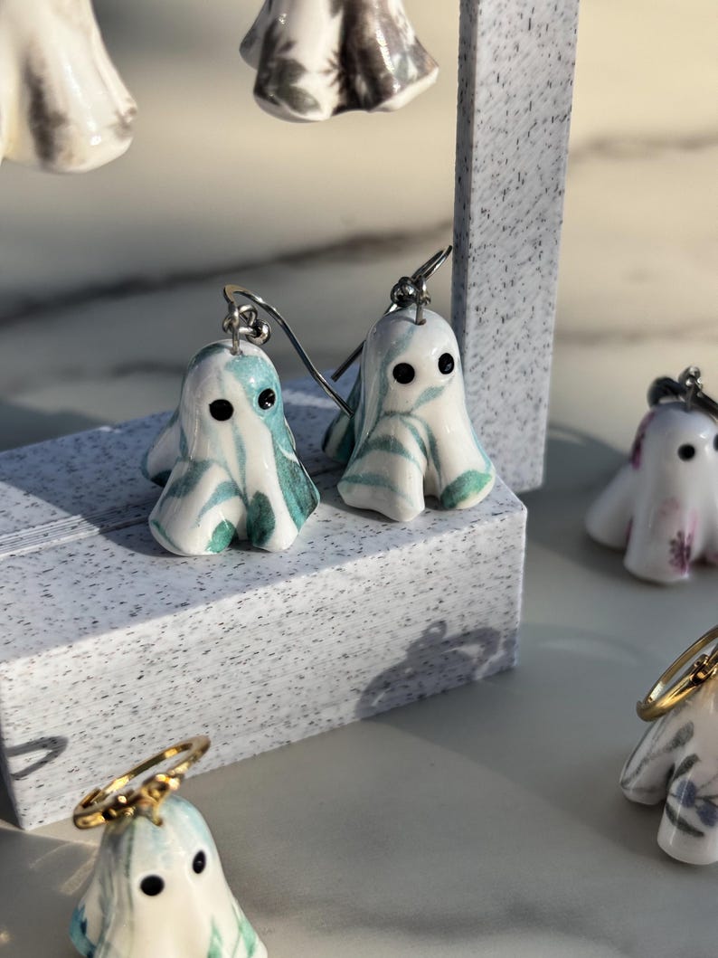 May include: A pair of drop earrings featuring small, ghost-shaped charms made of white ceramic with swirling teal and green marbled patterns. Each ghost has two small black dots for eyes and is attached to a silver-toned metal earring hook.