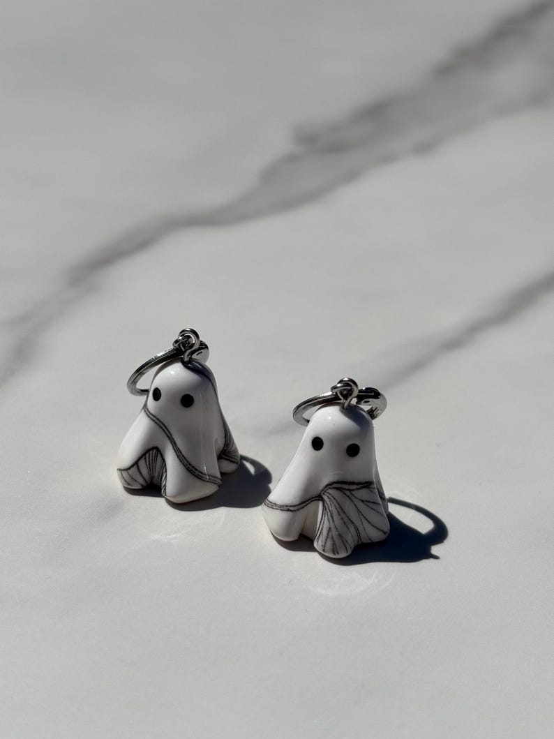May include: Two white ghost-shaped keychains with black eyes and gray shading. Each keychain has a silver-colored ring. The keychains are on a white surface with a marble pattern.