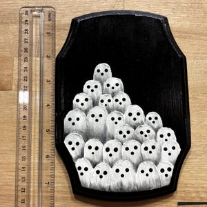 May include: A black wooden plaque featuring a painting of numerous white ghosts with black eyes. The plaque is approximately 8 inches tall and 6 inches wide. The ghosts are painted in a simple, cartoonish style, creating a spooky, Halloween-themed artwork.