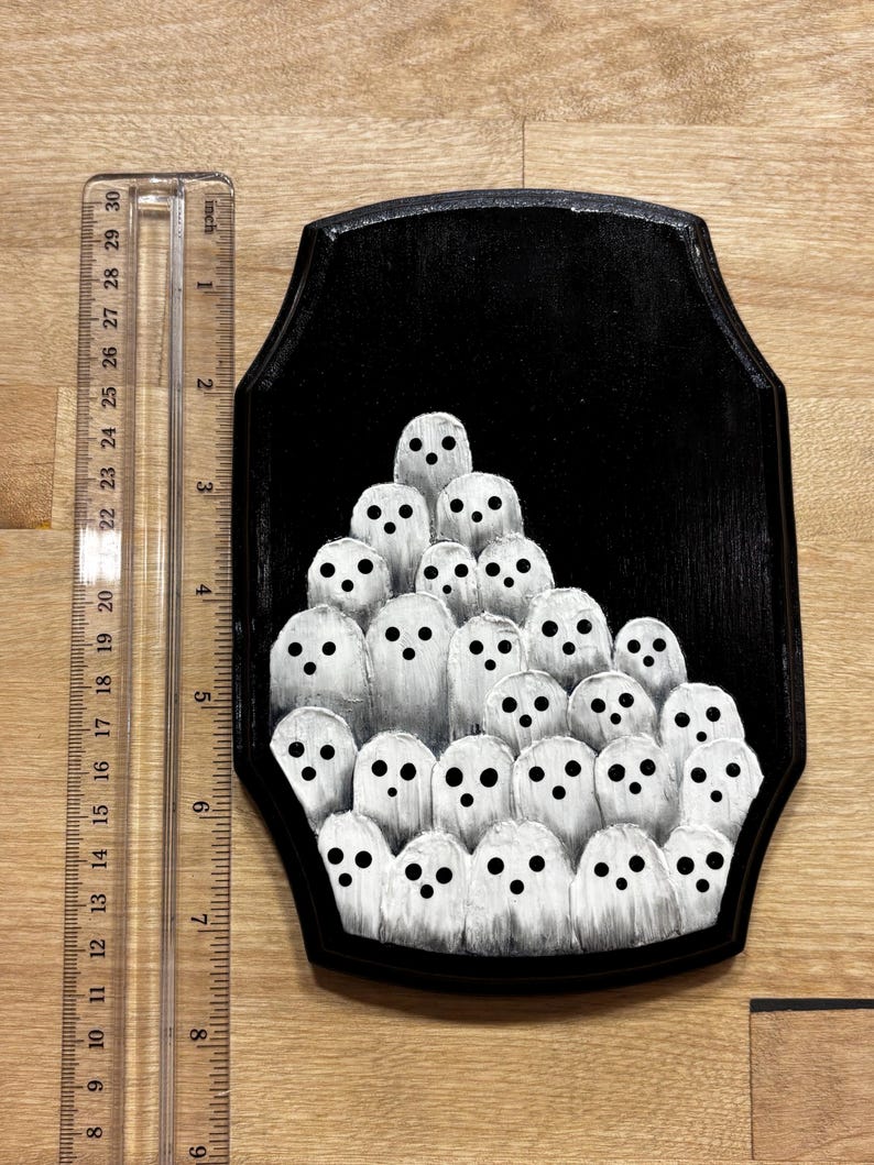 May include: A black wooden plaque featuring a painting of numerous white ghosts with black eyes. The plaque is approximately 8 inches tall and 6 inches wide. The ghosts are painted in a simple, cartoonish style, creating a spooky, Halloween-themed artwork.
