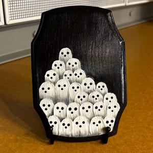 May include: A black wooden plaque featuring a hand-painted design of numerous white ghosts with black dot eyes. The ghosts are arranged in a cluster on the plaque, which is supported by a small black stand.