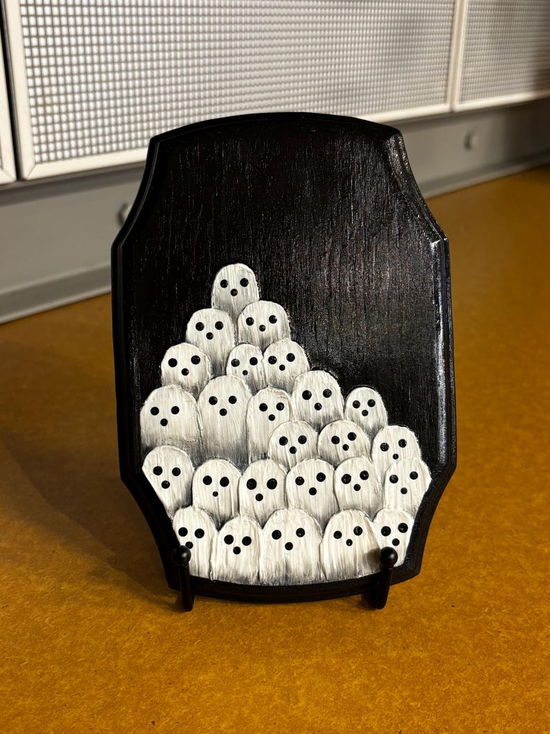 May include: A black wooden plaque featuring a hand-painted design of numerous white ghosts with black dot eyes. The ghosts are arranged in a cluster on the plaque, which is supported by a small black stand.