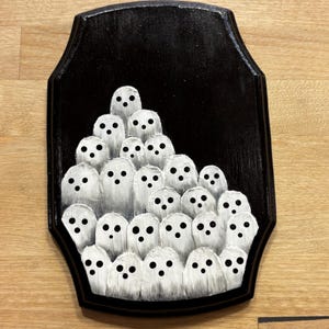 May include: A black wooden plaque featuring a group of white ghost figures with black dot eyes. The ghosts are painted with a textured, brushstroke effect, creating a spooky and whimsical Halloween decoration.