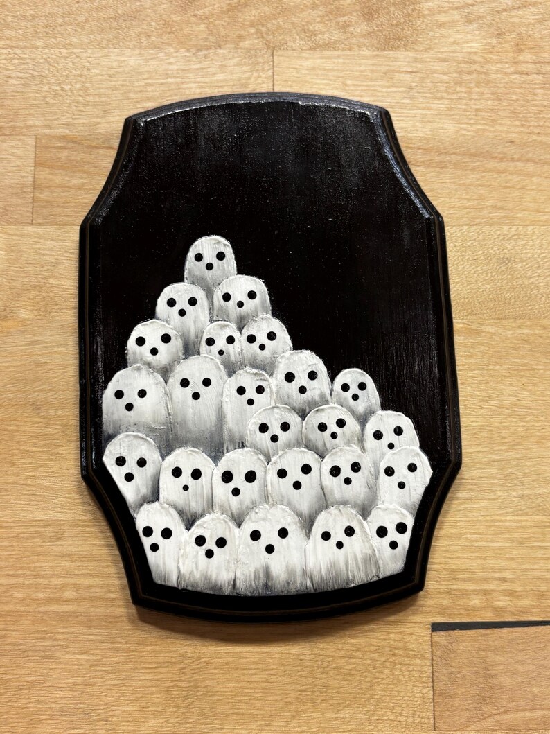 May include: A black wooden plaque featuring a group of white ghost figures with black dot eyes. The ghosts are painted with a textured, brushstroke effect, creating a spooky and whimsical Halloween decoration.