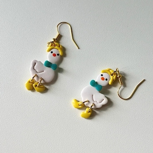 May include: A pair of whimsical goose earrings. Each earring features a white goose with yellow feet and a yellow hat. The goose has a teal bow tie and a gold-colored hook.