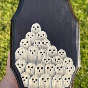 May include: A black wooden plaque featuring a gathering of white ghost figures with black dot eyes. The ghosts are arranged in a layered, clustered design. The plaque has a dark border and a textured surface.