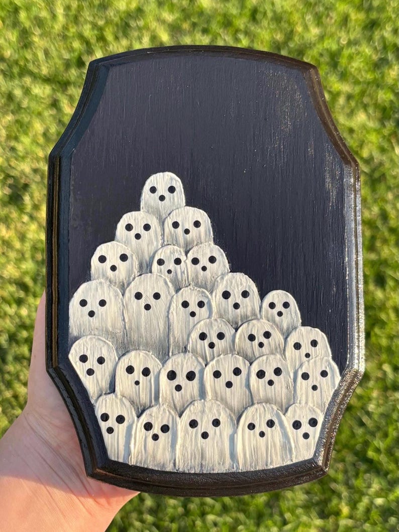 May include: A black wooden plaque featuring a gathering of white ghost figures with black dot eyes. The ghosts are arranged in a layered, clustered design. The plaque has a dark border and a textured surface.