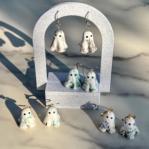 May include: Four pairs of ghost-shaped earrings featuring various floral and botanical patterns in shades of green, blue, and grey. Each ghost has two black dot eyes and is suspended from either silver-toned or gold-toned metal earring hooks.