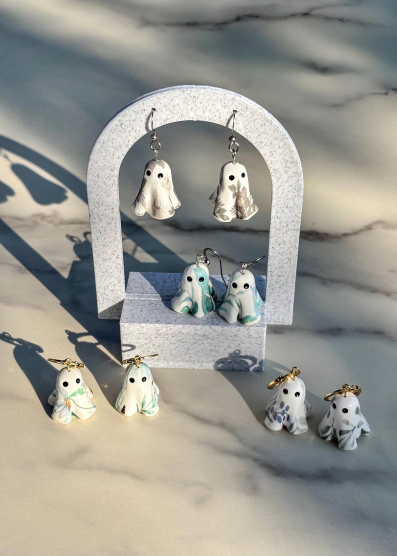 May include: Four pairs of ghost-shaped earrings featuring various floral and botanical patterns in shades of green, blue, and grey. Each ghost has two black dot eyes and is suspended from either silver-toned or gold-toned metal earring hooks.