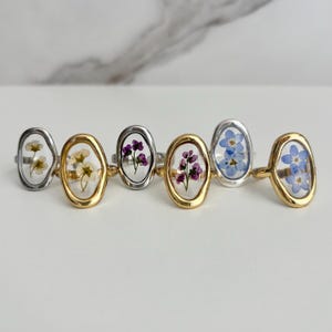 May include: A collection of six rings with oval-shaped clear resin settings, each containing dried flowers. The rings have either gold or silver bands. The flowers are various colors, including white, yellow, purple, and blue.