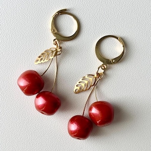 May include: A pair of cherry earrings. Each earring features two glossy red cherries with a gold-colored stem and a gold leaf accent. The earrings have gold-colored lever-back closures.