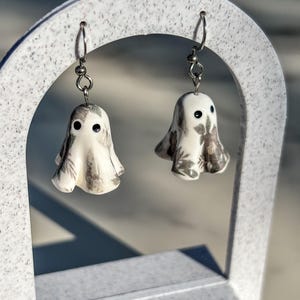 May include: A pair of drop earrings featuring small, white, ghost-shaped charms. Each ghost has two black dot eyes and a subtle, grey, leafy botanical pattern printed across its surface. The charms hang from silver-toned metal earring hooks.