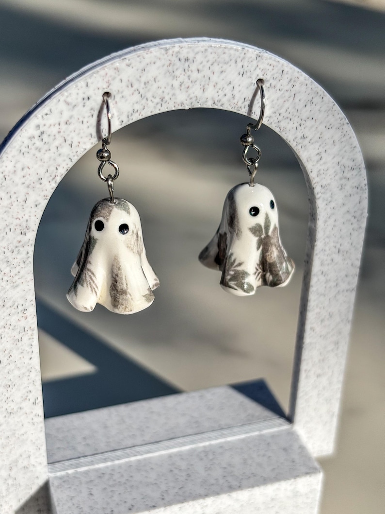 May include: A pair of drop earrings featuring small, white, ghost-shaped charms. Each ghost has two black dot eyes and a subtle, grey, leafy botanical pattern printed across its surface. The charms hang from silver-toned metal earring hooks.