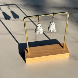 May include: A pair of drop earrings featuring white ghost-shaped charms with black circular eyes and green leaf patterns. The ghost charms hang from small gold-toned metal hoops. The earrings are suspended from a gold-toned metal display stand.