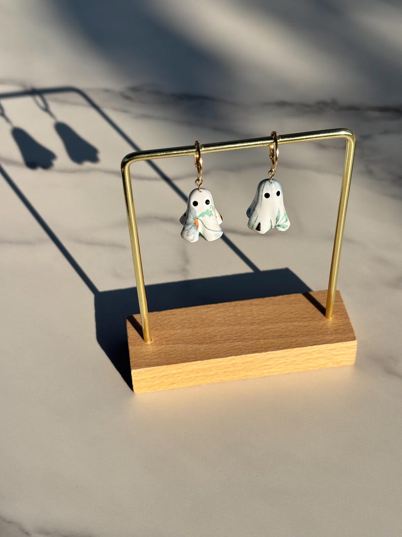 May include: A pair of drop earrings featuring white ghost-shaped charms with black circular eyes and green leaf patterns. The ghost charms hang from small gold-toned metal hoops. The earrings are suspended from a gold-toned metal display stand.