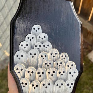May include: A black wooden plaque featuring a carved design of numerous white ghosts with black dot eyes. The ghosts are arranged in a layered, clustered pattern against a black background, creating a spooky, Halloween-themed decoration.