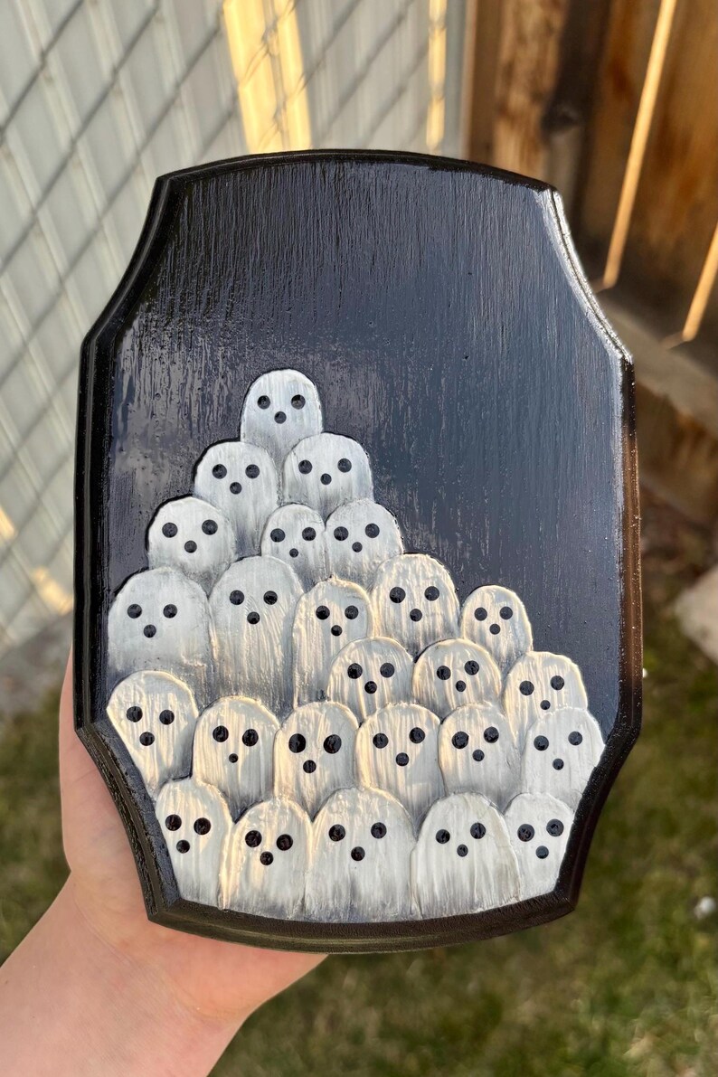 May include: A black wooden plaque featuring a carved design of numerous white ghosts with black dot eyes. The ghosts are arranged in a layered, clustered pattern against a black background, creating a spooky, Halloween-themed decoration.