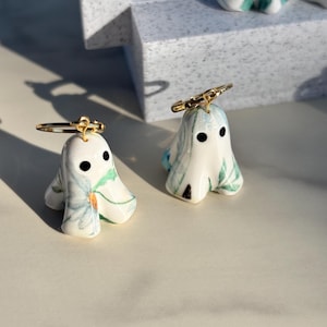May include: A pair of ghost-shaped earrings featuring a white ceramic base with blue and green floral patterns. Each ghost has two black circular eyes and is attached to a thin gold-toned metal hoop ring at the top of the head for hanging.
