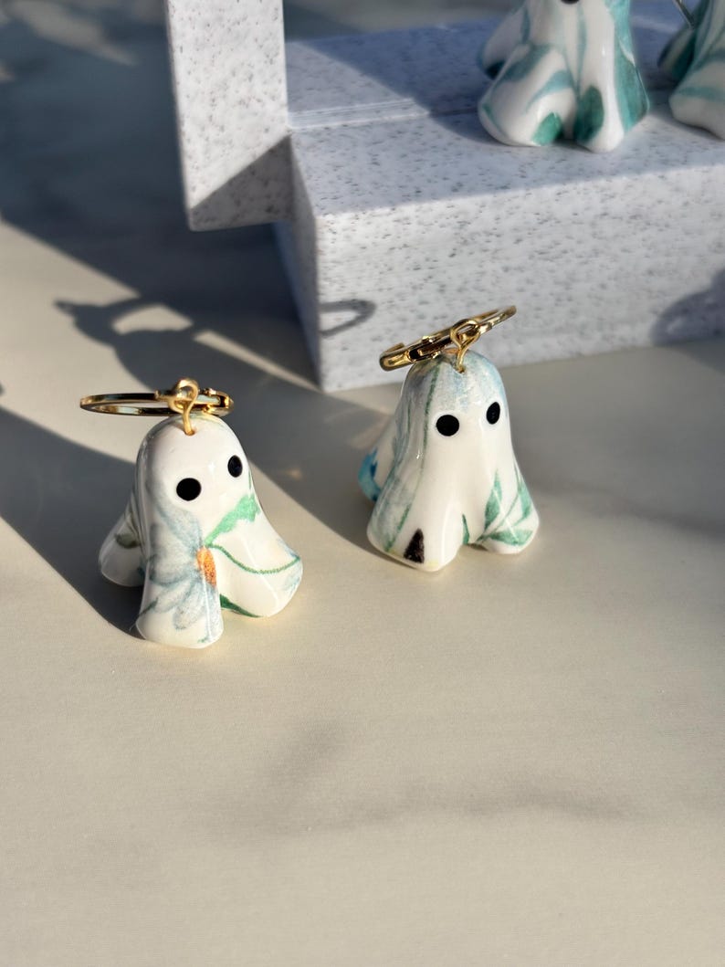 May include: A pair of ghost-shaped earrings featuring a white ceramic base with blue and green floral patterns. Each ghost has two black circular eyes and is attached to a thin gold-toned metal hoop ring at the top of the head for hanging.