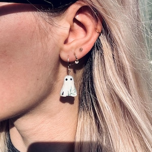 May include: A white ghost-shaped earring charm featuring two black circular eyes and a subtle green leaf pattern on the front. The charm is suspended from a thin gold-toned metal hoop earring that is secured through the earlobe.
