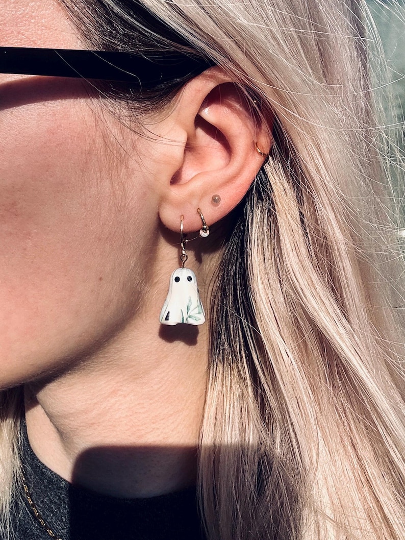May include: A white ghost-shaped earring charm featuring two black circular eyes and a subtle green leaf pattern on the front. The charm is suspended from a thin gold-toned metal hoop earring that is secured through the earlobe.