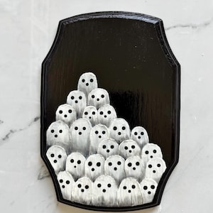May include: A black, shield-shaped wooden plaque featuring a cluster of white ghost figures with black dot eyes. The ghosts are painted with a textured, brushstroke effect, creating a spooky, Halloween-themed decoration.
