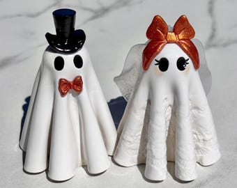 May include: Two white ghost figurines. The left ghost wears a black top hat and a metallic orange bow tie. The right ghost features a metallic orange bow on its head and a translucent white veil, with black eyes and eyelashes painted on its face.
