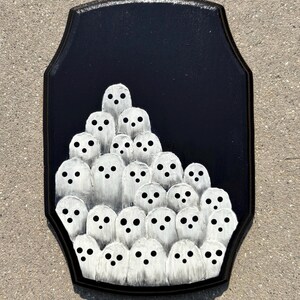 May include: A black wooden plaque featuring a gathering of white ghost figures with black dot eyes. The ghosts are clustered together against a black background, creating a spooky Halloween decoration.