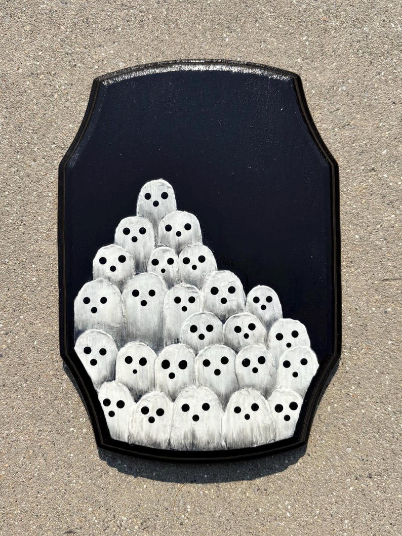 May include: A black wooden plaque featuring a gathering of white ghost figures with black dot eyes. The ghosts are clustered together against a black background, creating a spooky Halloween decoration.