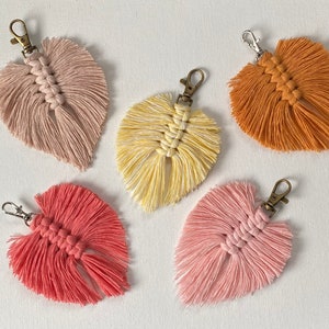 May include: Five colorful macrame leaf keychains with gold metal rings. The keychains are in shades of pink, peach, yellow, and orange.