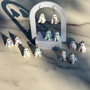 May include: A collection of small ghost-shaped earrings featuring various marbled and floral patterns in shades of white, grey, blue, green, and pink. Each ghost has two black dot eyes and is attached to a metal earring hook or hoop finding.