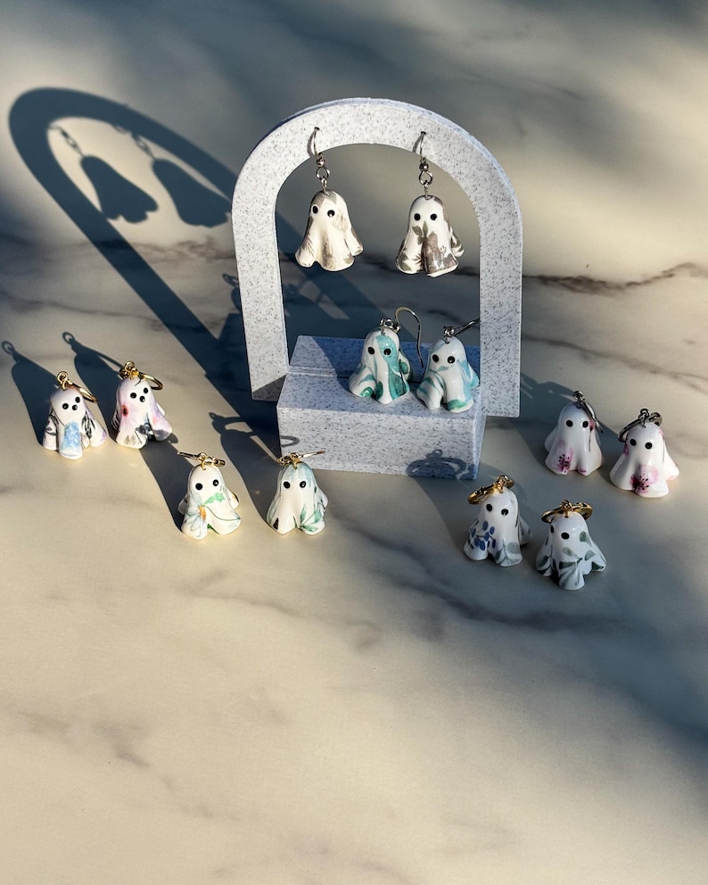 May include: A collection of small ghost-shaped earrings featuring various marbled and floral patterns in shades of white, grey, blue, green, and pink. Each ghost has two black dot eyes and is attached to a metal earring hook or hoop finding.