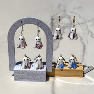 May include: Four pairs of ghost-shaped earrings hanging on two display stands. The ghosts are white with black eyes and feature various floral patterns in shades of pink, purple, and blue. Each ghost charm is attached to a metallic hoop earring.