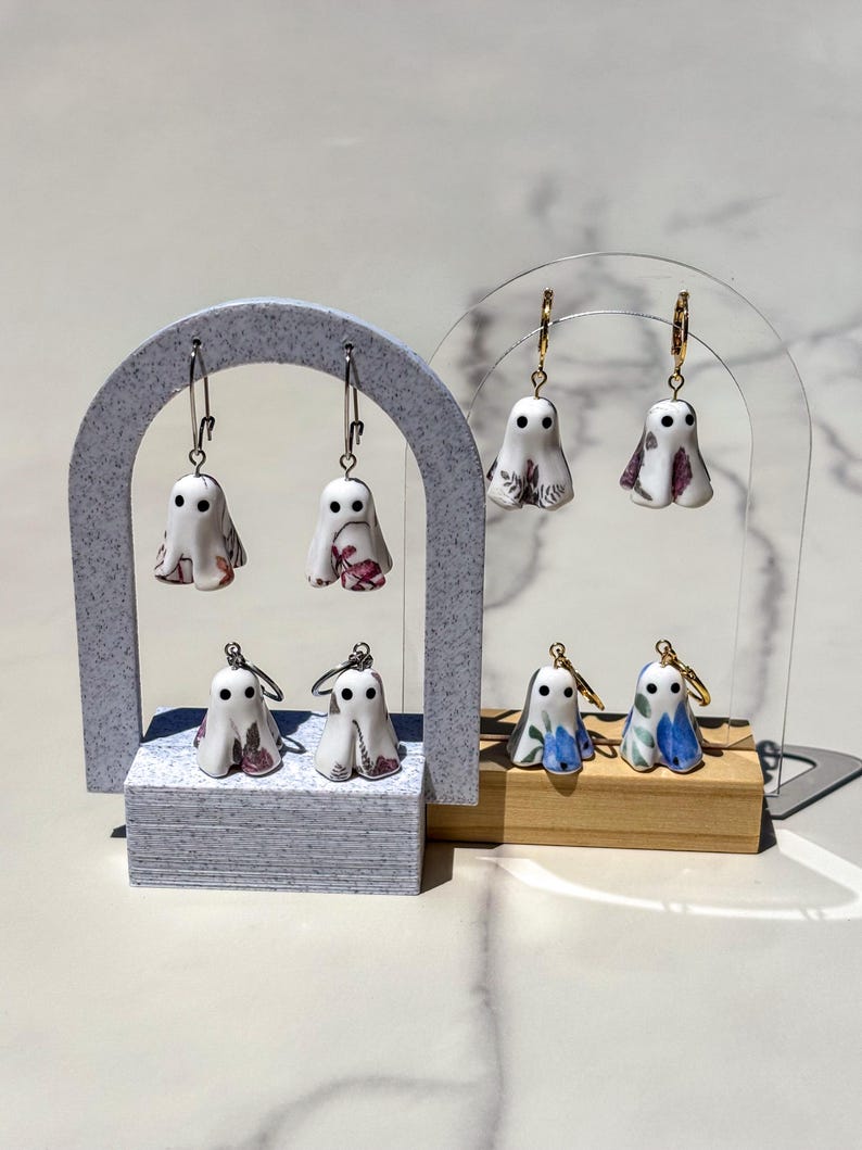 May include: Four pairs of ghost-shaped earrings hanging on two display stands. The ghosts are white with black eyes and feature various floral patterns in shades of pink, purple, and blue. Each ghost charm is attached to a metallic hoop earring.
