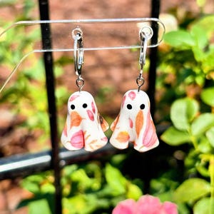 May include: A pair of dangle earrings featuring white ghost-shaped charms with a colourful autumn leaf pattern in shades of orange, pink, and brown. Each ghost has two small black circular eyes and hangs from a silver-toned metal hoop earring.