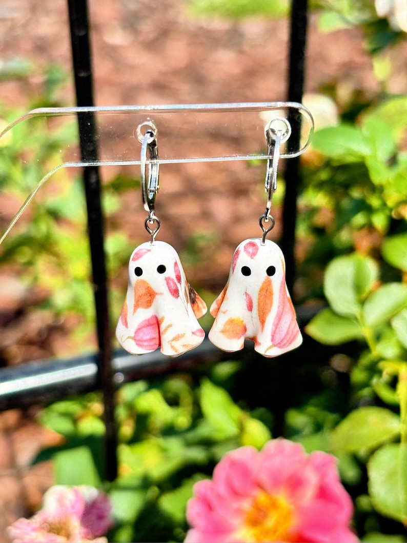 May include: A pair of dangle earrings featuring white ghost-shaped charms with a colourful autumn leaf pattern in shades of orange, pink, and brown. Each ghost has two small black circular eyes and hangs from a silver-toned metal hoop earring.